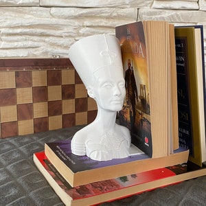 May include: White bust sculpture of an Egyptian queen with a tall headdress, used as a bookend. The bust is resting on a stack of books, including one with the title "As I  Est  Legenda".