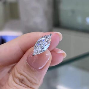 Op de afbeelding: Een marquise-geslepen diamant, met een briljante, langwerpige vorm. De diamant is helder en facetgeslepen, waardoor het licht wordt gereflecteerd. De zetting is van delicaat, zilverkleurig metaal. Dit beeld toont een sieraad.