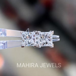 Op de afbeelding: Een paar oorknopjes met diamanten in prinsessenslijpsel, voorzien van heldere, gefacetteerde stenen in een metalen zetting met vier pootjes. De oorbellen worden vastgehouden door een transparante plastic pincet, met de tekst MAHIRA JEWELS.