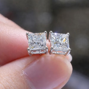 Op de afbeelding: Een paar oorknopjes met vierkante, princess-geslepen heldere stenen in zilverkleurige metalen zettingen met vier pootjes. De stenen hebben meerdere facetten die het licht weerkaatsen, wat zorgt voor een heldere en sprankelende uitstraling.