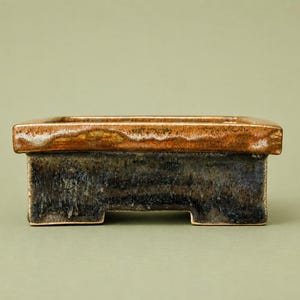 May include: A rectangular ceramic bonsai pot featuring a dark, mottled black and grey base with a contrasting smooth, glossy amber-brown rim. The vessel has a wide, flat top edge and is supported by four small, integrated feet at the corners.