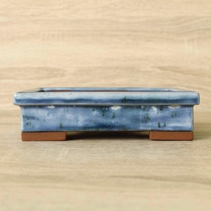 May include: A rectangular ceramic bonsai pot featuring a glossy blue glaze with subtle white and dark speckles. The pot has a thick, flat rim and is supported by four small, unglazed terracotta-coloured feet at the base, showing a smooth finish.