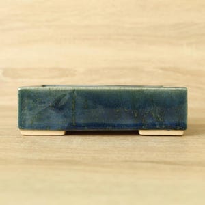Peut inclure: Un pot de fleurs rectangulaire en céramique bleu foncé avec une finition brillante. Le pot a un profil bas, reposant sur deux petits pieds blanc cassé. Le fond est un grain de bois clair.