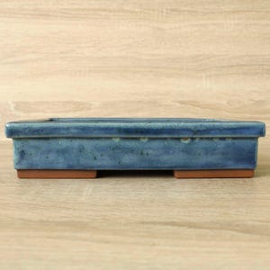 May include: A rectangular ceramic bonsai pot featuring a glossy blue glaze with subtle variations in tone. The pot has a flat rim and is supported by four small, unglazed terracotta-colored feet at the base, providing a stable foundation.