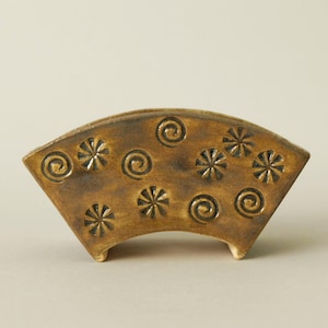 Op de afbeelding: Een bruin, keramisch decoratief stuk in de vorm van een waaier, versierd met reliëfspiraal- en sterpatronen. Het object heeft een gebogen basis en is waarschijnlijk een klein schaaltje of houder. Afmetingen in centimeters.