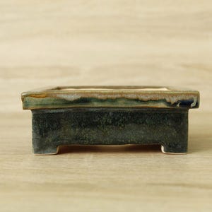 May include: A rectangular ceramic bonsai pot with a dark, mottled green and black glaze on the sides and a lighter, variegated green and beige glaze along the top rim. The vessel features four short, sturdy feet and a flat, open top surface.