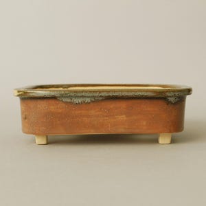 May include: A rectangular ceramic bonsai pot with a matte terracotta-coloured exterior and a smooth, cream-coloured interior. The rim features a dark, mottled green glaze that drips slightly down the sides. The pot stands on four small, light-coloured feet.