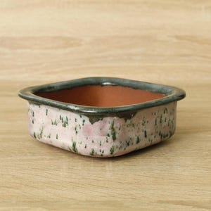Puede incluir: Una jardinera cuadrada de cerámica con un exterior moteado rosa y verde y un borde oscuro. El interior es de color marrón rojizo. La jardinera está sobre una superficie de madera marrón claro.