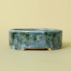 May include: A shallow, round ceramic bonsai pot with a glossy, mottled glaze in shades of sage green, blue, and grey. The vessel features a smooth, slightly uneven rim and rests on four small, integrated feet that elevate the base slightly.