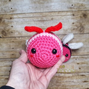 May include: Two crocheted bee plushies, one pink with white stripes and red antennae, the other black and yellow with white wings. The pink bee is being held in a hand.