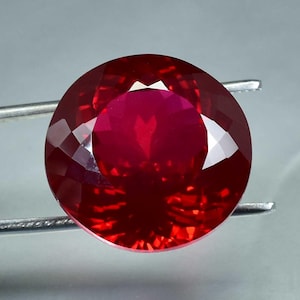 May include: A deep red gemstone with a round cut and a faceted surface. The gemstone is set in a silver setting.