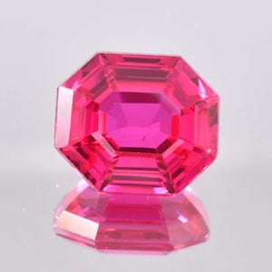May include: A faceted, octagonal-cut gemstone in a vibrant shade of pink. The gem's multiple facets reflect light, creating a brilliant display. The gem is set against a reflective surface, enhancing its color and shape.