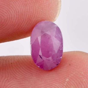 May include: Oval-cut, faceted ruby gemstone. The ruby is a vibrant pinkish-red color, with visible internal inclusions. The gem is held between two fingers against a white background, showcasing its polished facets and natural characteristics.