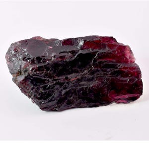 May include: A raw, uncut gemstone with a deep burgundy color. The surface of the stone has a rough, textured appearance with areas of high gloss. The gemstone is set against a white background.