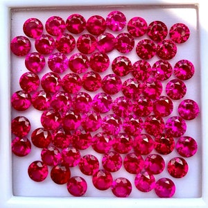 May include: A collection of loose ruby red gemstones, displayed in a white square box. The gemstones are various sizes and shapes, with a polished finish.