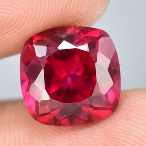 May include: A faceted, cushion-cut ruby gemstone. The gem is a deep, saturated red color with multiple facets that reflect light. The ruby is held between two fingers, showcasing its size and brilliance.