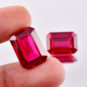 May include: Two vibrant red emerald cut gemstones. The larger gemstone is being held by a finger, while the smaller gemstone is resting on a white surface.