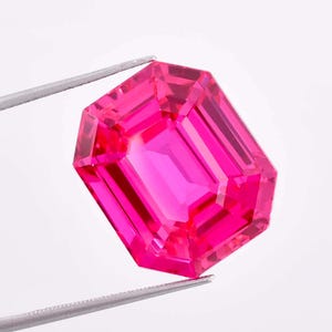 May include: A vibrant, faceted, emerald-cut gemstone in a vivid pink hue. The gem is held by silver tweezers against a white background, showcasing its clarity and brilliance. The gemstone's rectangular shape and step-cut facets are clearly visible.