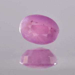 Puede incluir: Una gema rosa facetada, de forma ovalada, con una superficie reflectante. La gema es de un tono rosa claro con facetas internas que captan la luz. La gema se refleja en la superficie inferior.