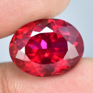 Peut inclure: Une grande pierre précieuse rouge de forme ovale avec une taille facetée. La pierre précieuse est tenue dans une main, et la lumière se reflète sur les facettes, créant un éclat.