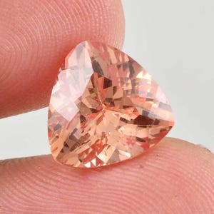 May include: A faceted, peach-colored gemstone with a triangular shape. The gem has multiple facets that reflect light, creating a sparkling effect. The stone is held between two fingers, showcasing its size and color.