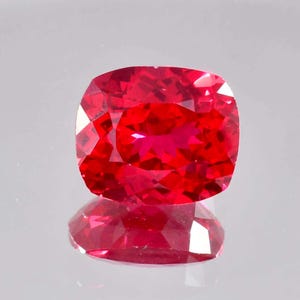 May include: A faceted, cushion-cut, red gemstone. The gem is a vibrant, deep red color with multiple facets that reflect light. The gemstone is displayed on a reflective surface, creating a mirror image.