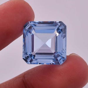 May include: A light blue gemstone with a square emerald cut. The gemstone is faceted and has a high degree of clarity.