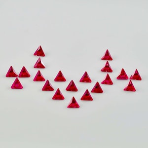 May include: A collection of twenty ruby red triangular gemstones arranged in a heart shape on a white background.