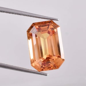 May include: A faceted, rectangular gemstone with an emerald cut. The gem is held by silver tweezers. The stone has a warm, orange-yellow color with internal facets that reflect light.