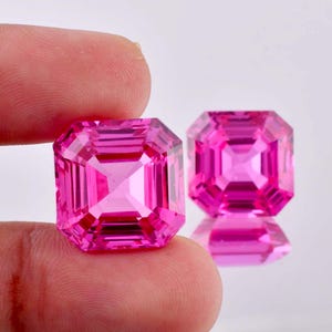 May include: Two pink square-shaped gemstones with a faceted cut. The gemstones are held in a hand.