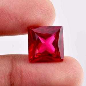 May include: A square-cut, faceted, bright red gemstone. The gem has a cross-like pattern in the center, with sharp, clean edges. The gemstone is held between two fingers.