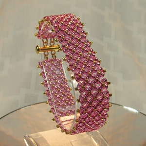 May include: A pink and gold beaded bracelet with a clasp closure. The bracelet is made with small, round beads that are arranged in a geometric pattern.