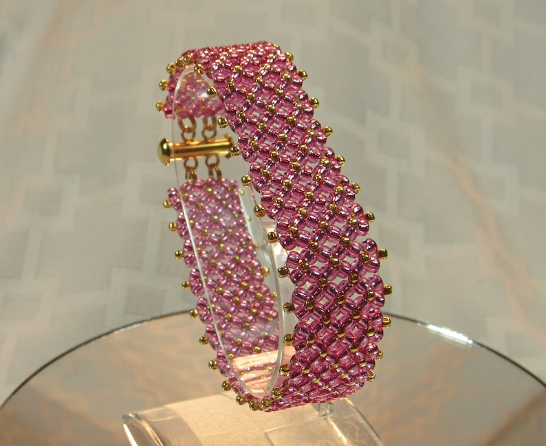 May include: A pink and gold beaded bracelet with a clasp closure. The bracelet is made with small, round beads that are arranged in a geometric pattern.