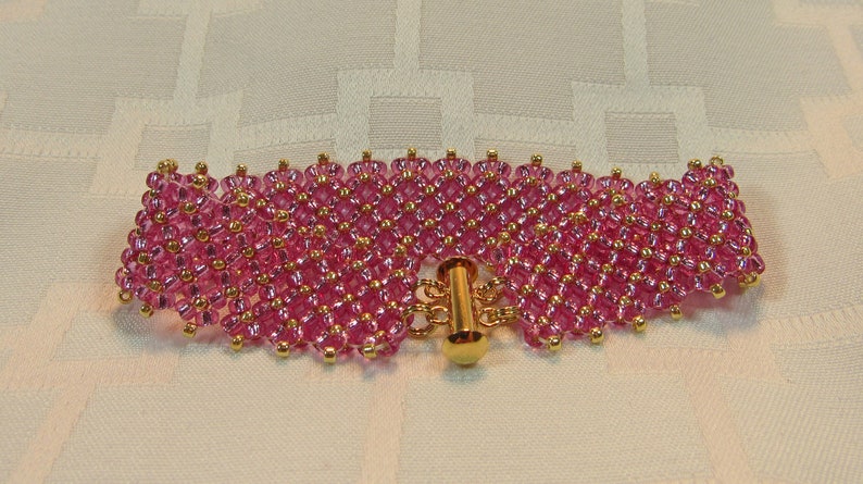May include: A pink beaded bracelet with a gold clasp. The bracelet is made of small, round beads that are arranged in a pattern. The clasp is a simple, gold-toned design.