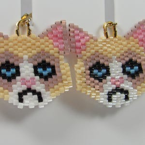 May include: A pair of beaded cat face earrings. The earrings feature a light yellow, pink, white, and black color scheme. The cat faces have blue eyes and a sad expression. The earrings have gold-colored hooks.