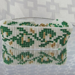 Könnte beinhalten: Ein weißes Perlenarmband mit einem grünen und goldenen Blumenmuster. Das Armband ist aus kleinen, runden Perlen gefertigt und hat ein schlichtes, elegantes Design.