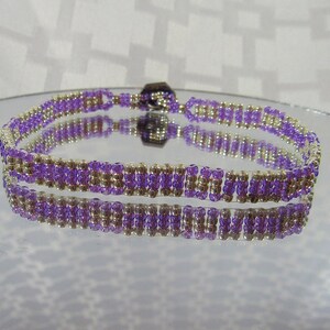 May include: A beaded bracelet with a repeating pattern of purple and gold beads. The bracelet is displayed on a reflective surface, showcasing its symmetrical design and color contrast. The bracelet is approximately 0.5 inches wide.