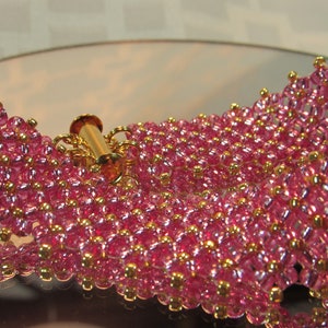 May include: A pink and gold beaded bracelet with a gold clasp. The bracelet is made of small, round beads that are arranged in a pattern. The beads are iridescent and have a slight shimmer to them.
