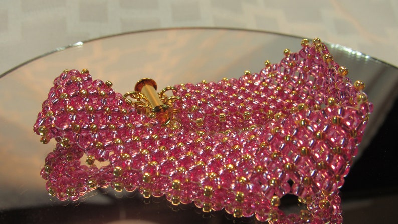May include: A pink and gold beaded bracelet with a gold clasp. The bracelet is made of small, round beads that are arranged in a pattern. The beads are iridescent and have a slight shimmer to them.