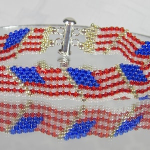 Puede incluir: Una pulsera de cuentas con un diseño que recuerda a la bandera estadounidense. La pulsera está hecha con cuentas rojas, blancas y azules, con un cierre plateado. El diseño incluye cuadrados azules con estrellas blancas y rayas rojas y blancas.