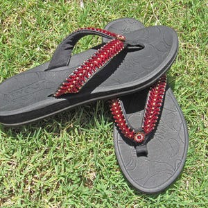 May include: A pair of black flip-flops featuring a thick sole with a subtle embossed paisley pattern. The Y-shaped straps are embellished with rows of small, vibrant red beads and a circular red beaded accent at the center of each toe post.