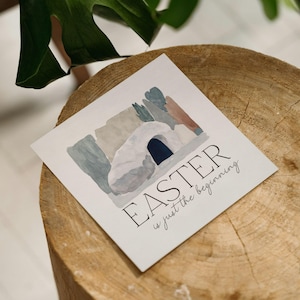 May include: A square Easter card featuring a watercolor illustration of a tomb with a dark blue entrance. The card has the text "EASTER is just the beginning" in black script. The card is on a wooden surface.