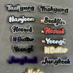 May include: A collection of glittery hair clips with names like "Taehyung", "Namjoon", and "Jimin" in various colors. The clips have a cursive font and are set against a gray background.