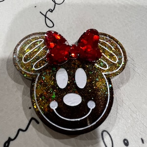 May include: A gingerbread-themed resin pin with a red glitter bow, white eyes, and a smiling mouth. The pin is decorated with gold glitter.