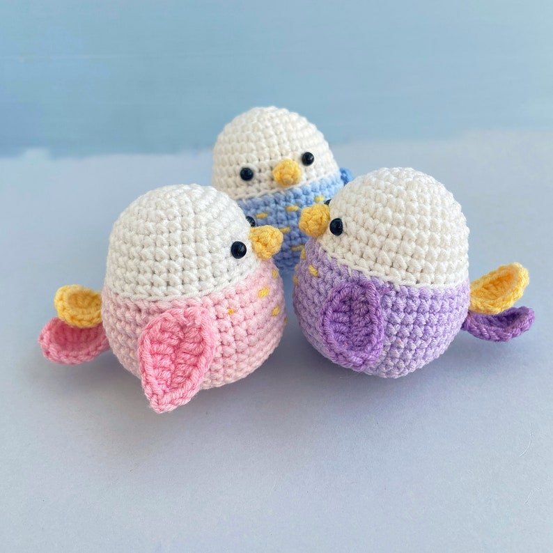 May include: Three handmade crochet bird toys in pastel colors. The birds have white heads, black eyes, yellow beaks, and colorful bodies. One bird is pink, another is purple, and the third is wearing a blue scarf. The wings are yellow and pink.
