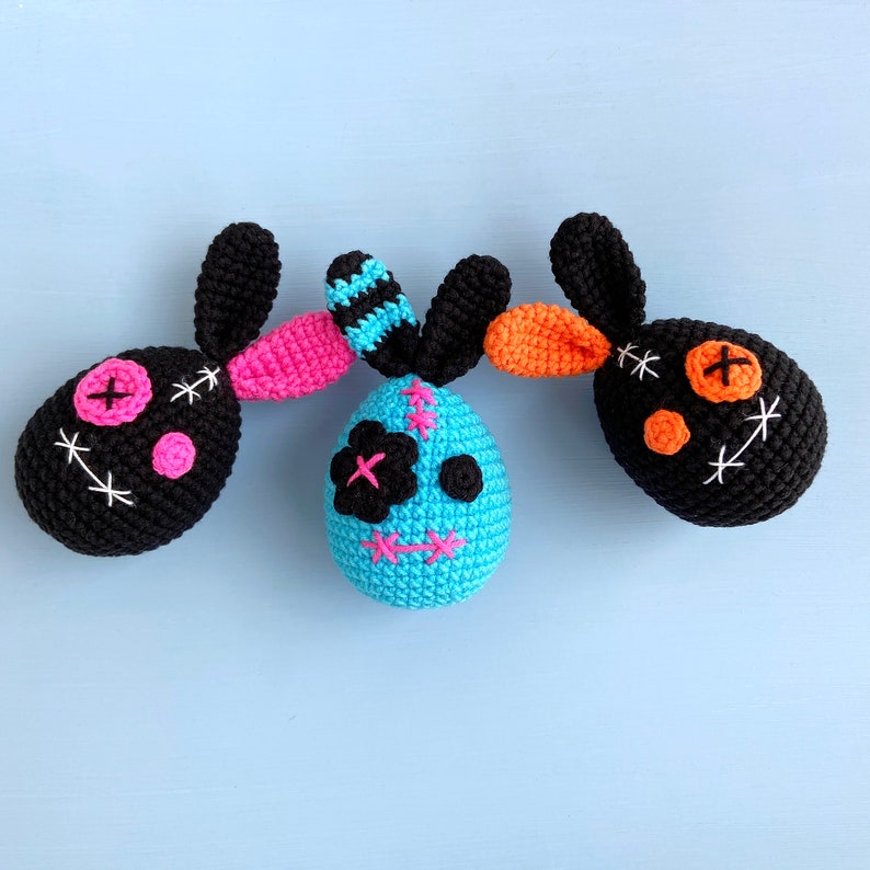 May include: Three crocheted bunny plushies with different coloured faces. The first bunny is black with white stitching and pink accents. The second bunny is blue with white stitching and pink accents. The third bunny is black with orange and white stitching.