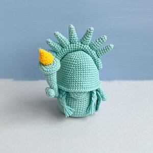 May include: A crocheted amigurumi doll of the Statue of Liberty, with a green body and a yellow torch. The doll is standing on a white surface.