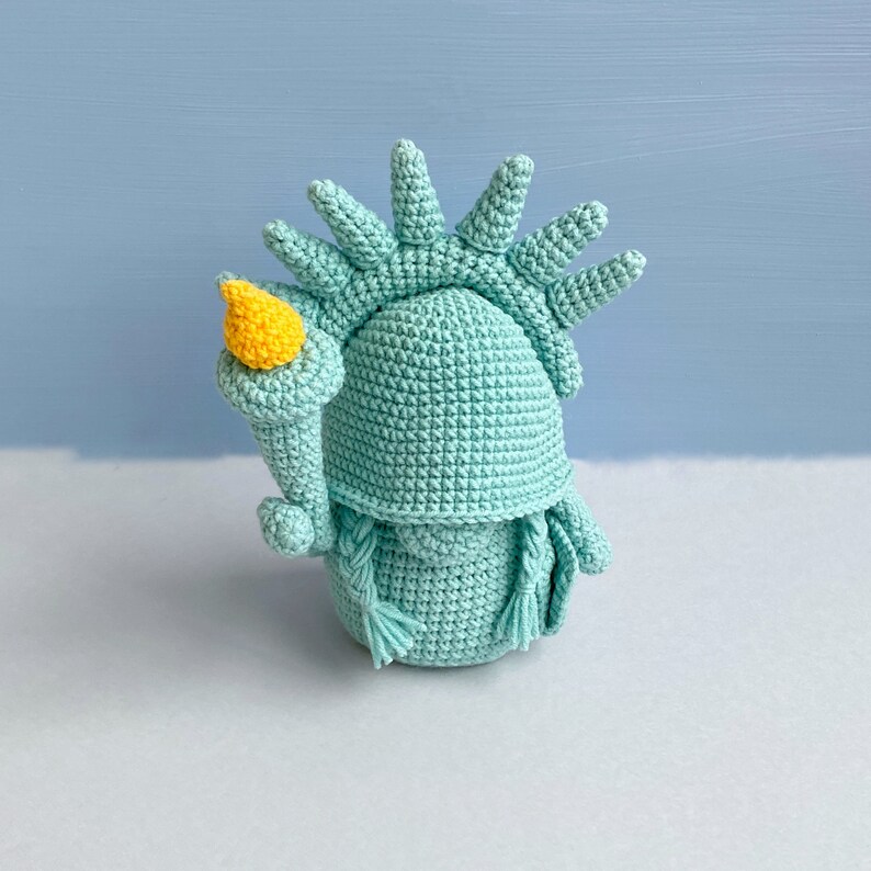 May include: A crocheted amigurumi doll of the Statue of Liberty, with a green body and a yellow torch. The doll is standing on a white surface.