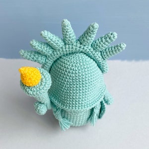 May include: A crocheted plush toy shaped like the Statue of Liberty. The toy is light blue with a yellow torch and a green base.