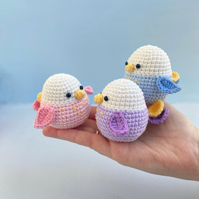 May include: Three handmade crochet bird figurines in pastel colors. Each bird has a white head, black eyes, and a yellow beak. The bodies are pink, purple, and blue, with matching wings and feet. The birds are held in a hand against a light blue background.